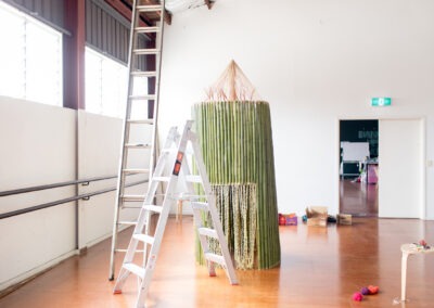photo of cut standing bamboo constructed in a round shape with pointed wool roof to be a art installation, within a dance space studio. Their are 2 ladders and things around showing it's a work in progress