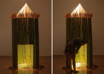 peering inside a round bamboo structure with a yellow wool door you can partially see through. The art installation is set up in a studio space at night with just one spotlight above lighting the structure.