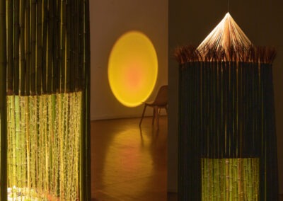 two close up photos put together of a round bamboo structure with a yellow wool door you can partially see through. The art installation is set up in a studio space at night with just one spotlight above lighting the structure and a chair and sunset light (projected onto a wall) to the right hand side