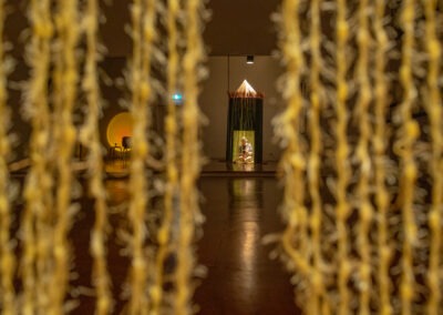 a shot looking out from inside a 2m bamboo art installation made into a round shelter with a front opening of thin yellow wool people can climb into. The photo is taken at night when the studio space is only lit by a small warm spotlight. The structure faces a mirror that reflects back.