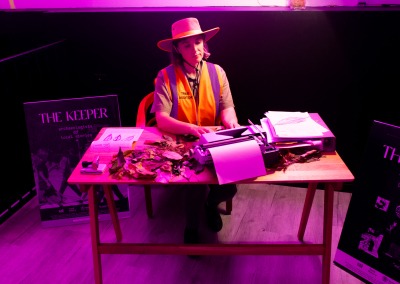 photo of a lady wearing hi vis behind a desk in an office. She appears to be typing on a typewriter with paper and leaves on the desk. The scene is lit but a magenta light