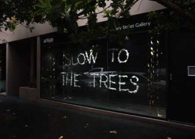 photo of a window art gallery. The window shows white lettering (spelt out in porcelain letters) 'SLOW TO THE TREES'.