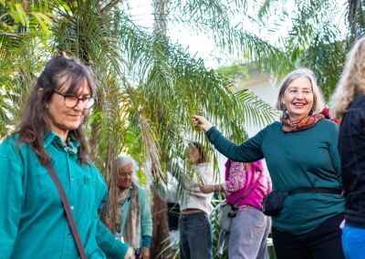 a lady smiling and touching leaves of a low palm tree, surrounded by other individuals in the frame