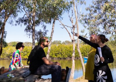 four inidviduals in front of a river, one is sitting on a park table, the other is pointing, all figures are blurry and the water and landscape behind are in focus
