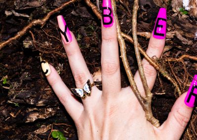 A photo of a left hand wearing fake nails with the word "cyber" spelt out across the nails. The hands sit on top of a woody forest floor.