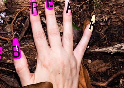 A photo of a right hand wearing fake nails with the word "break" spelt out across the nails. The hands sit on top of a woody forest floor.