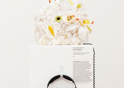 white handmade fabric flowers, appear on top of a plinth within a white walled gallery. The flowers are constructed into a cube. The plinth has a pair of black headphones hanging from a hook on the front of the plinth