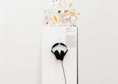 white handmade fabric flowers, appear on top of a plinth within a white walled gallery. The flowers are constructed into a cube. The plinth has a pair of black headphones hanging from a hook on the front of the plinth
