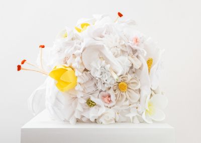 white and yellow handmade fabric flowers, appear on top of a plinth within a white walled gallery. The flowers are constructed into a cube.