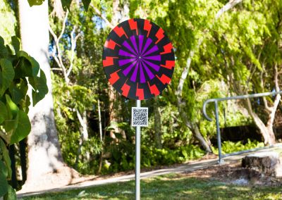 A photo taken outside at Noosa, that shows an art sign post in the middle. The post has a round purple, red and black psychedelic graphic and sits in a park next to a tree.
