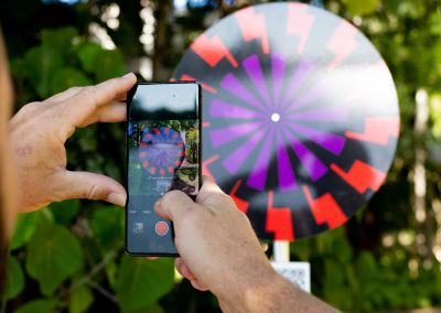 A photo taken outside at Noosa, that shows an art sign post in the middle. The post has a round purple, red and black psychedelic graphic and sits in a park next to a tree. The photo also shows someone (cropped) taking a photo of the QR code with their mobile phone