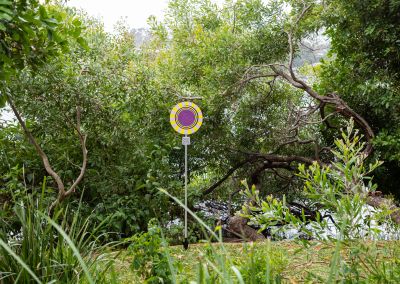 A photo taken outside at Noosa, that shows an art sign post in the middle. The post has a round purple, yellow and black psychedelic graphic and sits in a park in front of trees on the river.