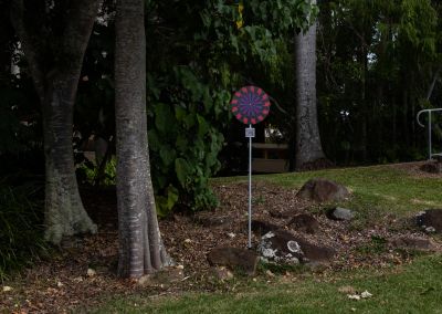 A photo taken outside at Noosa, that shows an art sign post in the middle. The post has a round purple, red and black psychedelic graphic and sits in a park next to a tree.